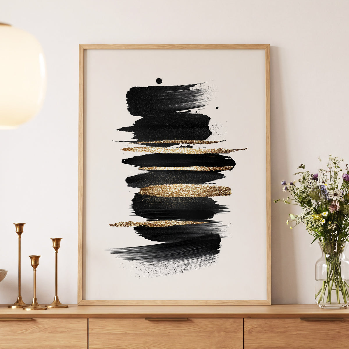 Black Gold Brushstrokes: a framed abstract art print featuring horizontal black brushstrokes with textured gold accents on a white background, displayed in a light wood frame on a wooden console table in a styled room setting.