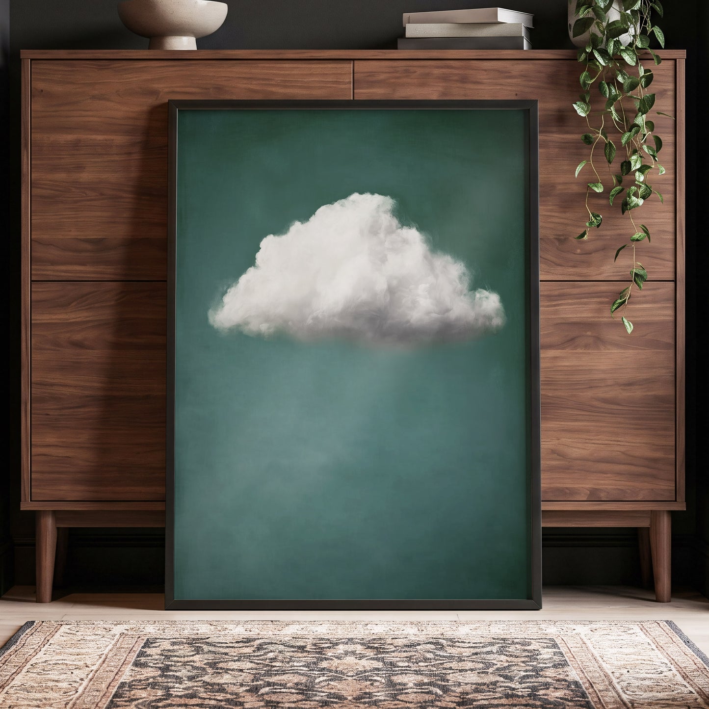 A framed vertical art print featuring a single fluffy white cloud against a deep teal-green background, leaning against a dark wood cabinet in a styled room with a hanging plant and patterned rug.