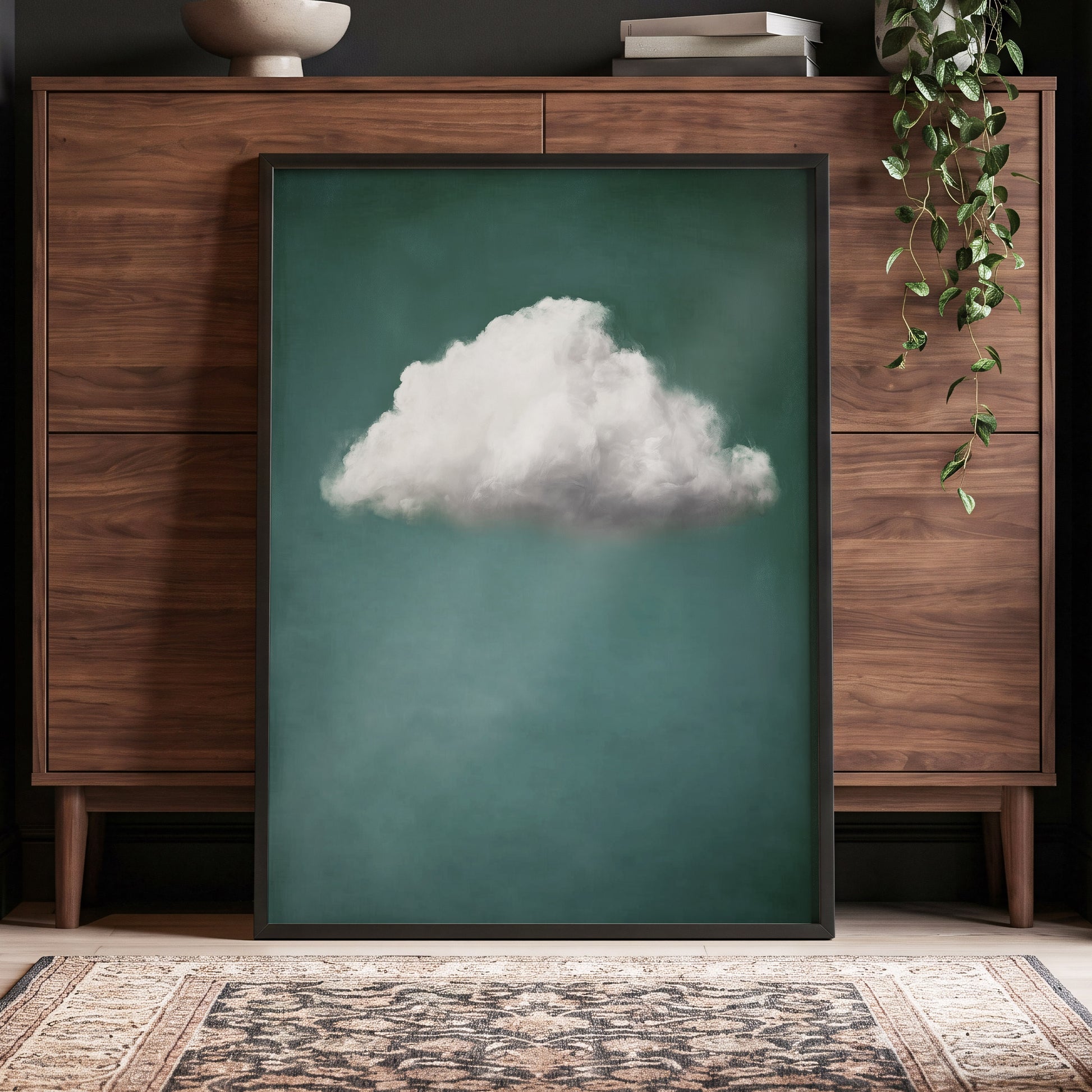 A framed vertical art print featuring a single fluffy white cloud against a deep teal-green background, leaning against a dark wood cabinet in a styled room with a hanging plant and patterned rug.
