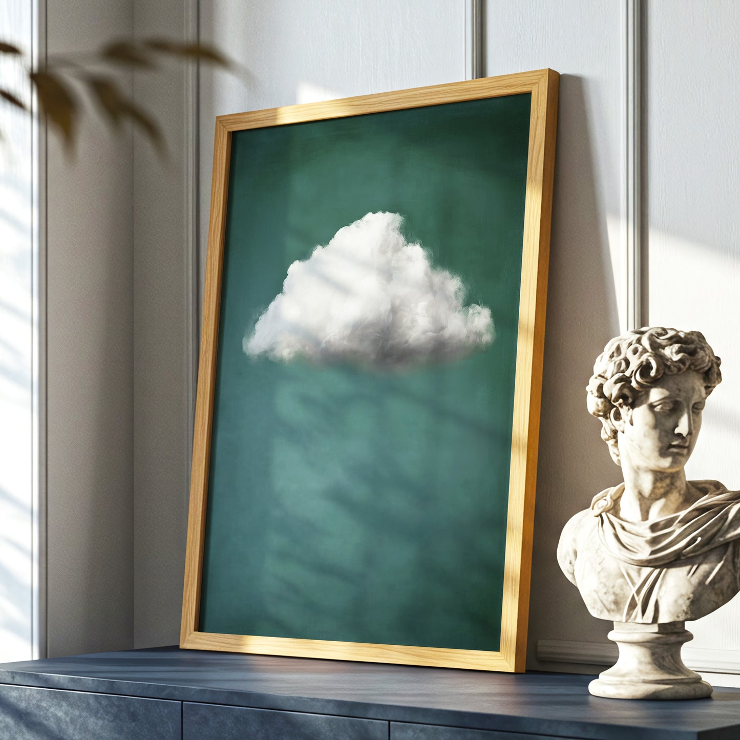 A framed wall art print leaning on a dark blue console table in a styled interior. The artwork shows a single white cloud against a deep teal-green background, enclosed in a light wood frame.