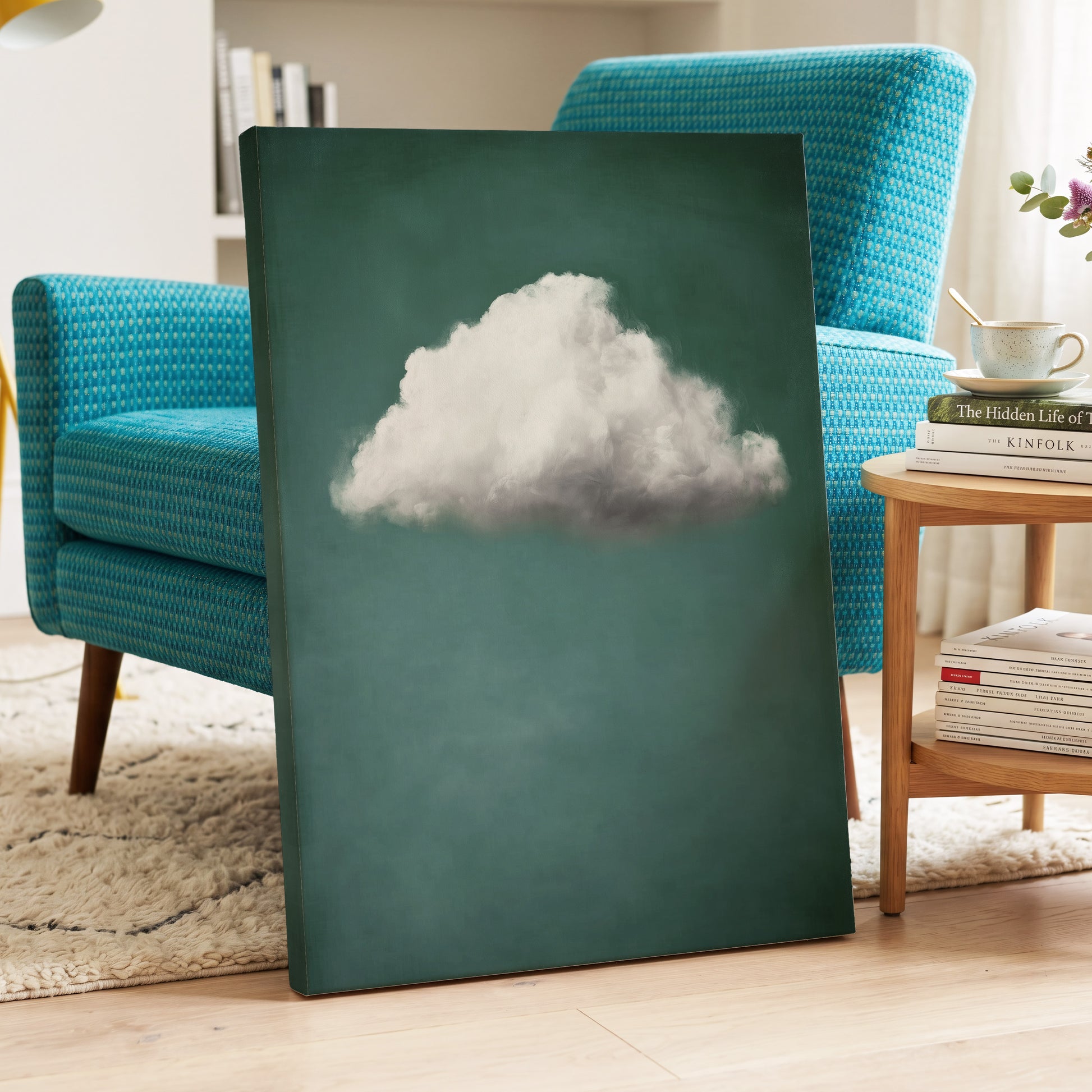 A vertical canvas wall art print leaning against a turquoise armchair in a living room setting. The artwork features a single fluffy white cloud against a dark teal-green background.