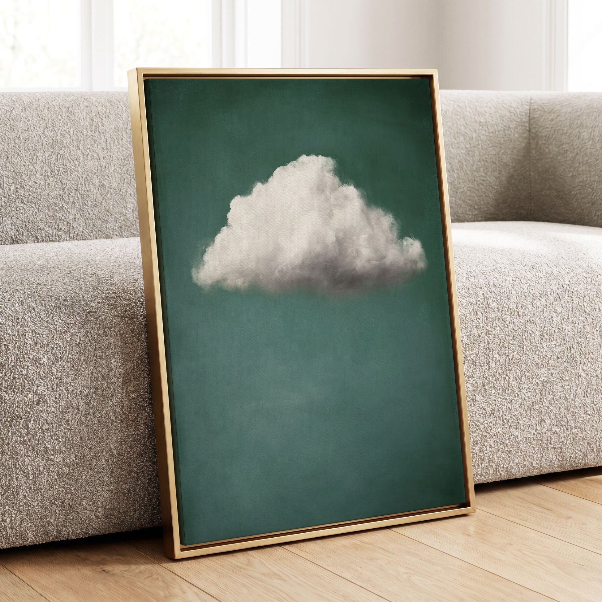 A framed art print featuring a single fluffy white cloud against a deep teal-green background, leaning against a textured beige sofa on a light wood floor.