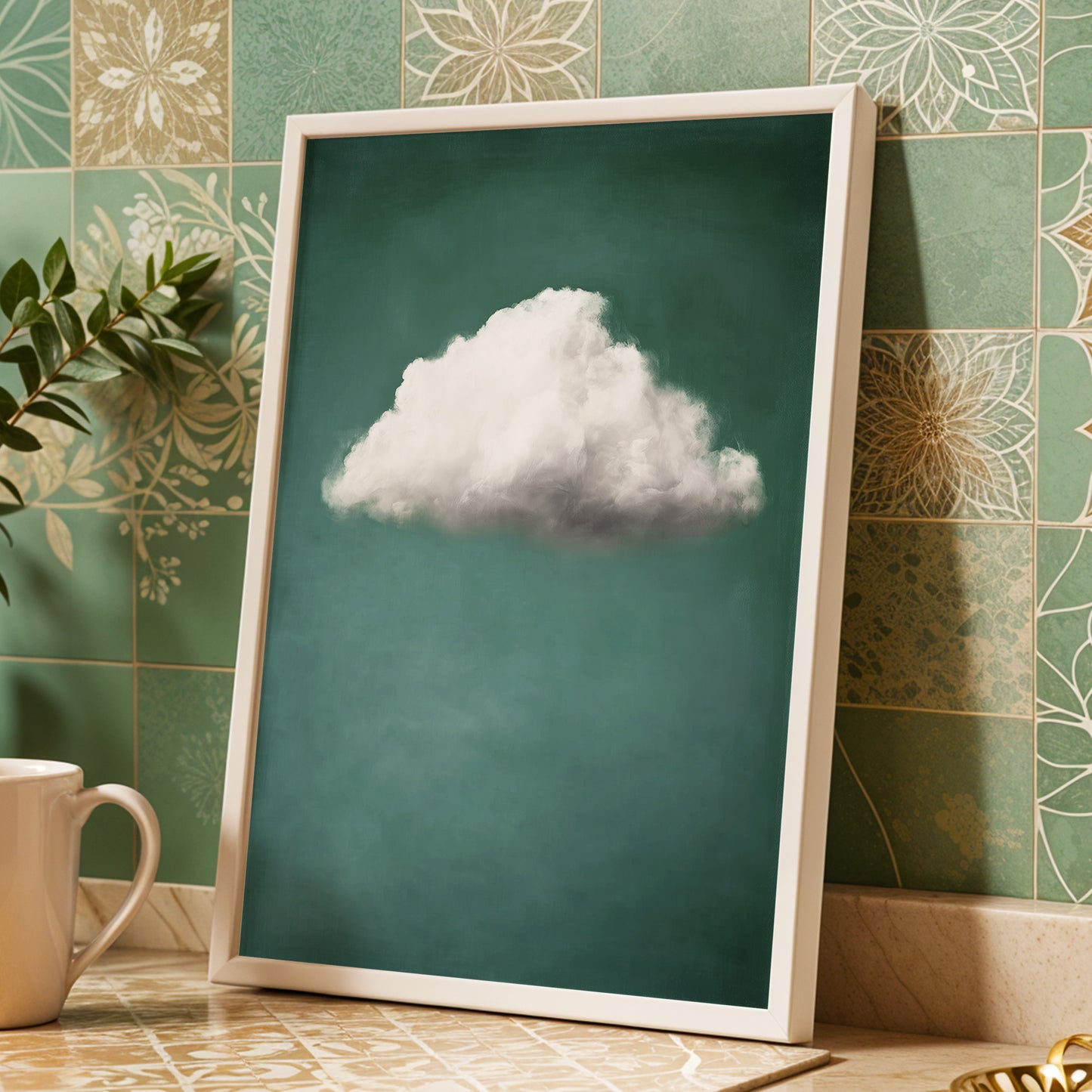 A framed art print featuring a single fluffy white cloud against a deep teal background, leaning against a green patterned tile wall on a countertop next to a white mug.