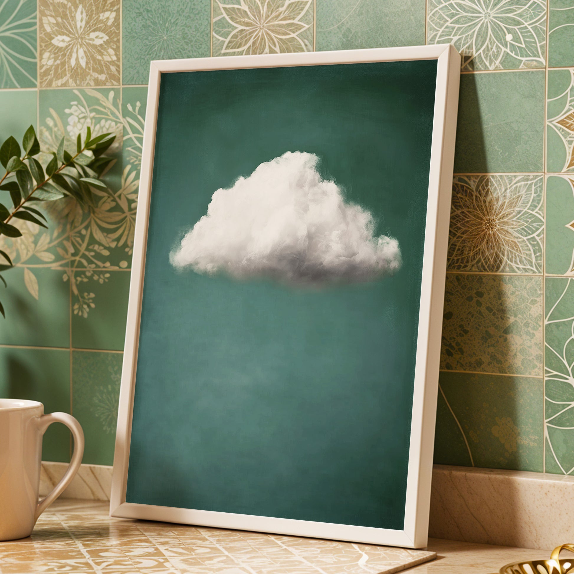 A framed art print featuring a single fluffy white cloud against a deep teal background, leaning against a green patterned tile wall on a countertop next to a white mug.