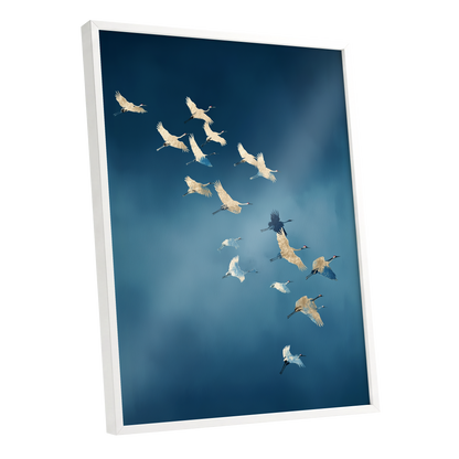 A framed wall art print showing a flock of cranes flying across a deep blue sky, photographed at an angle with a white frame and visible depth.