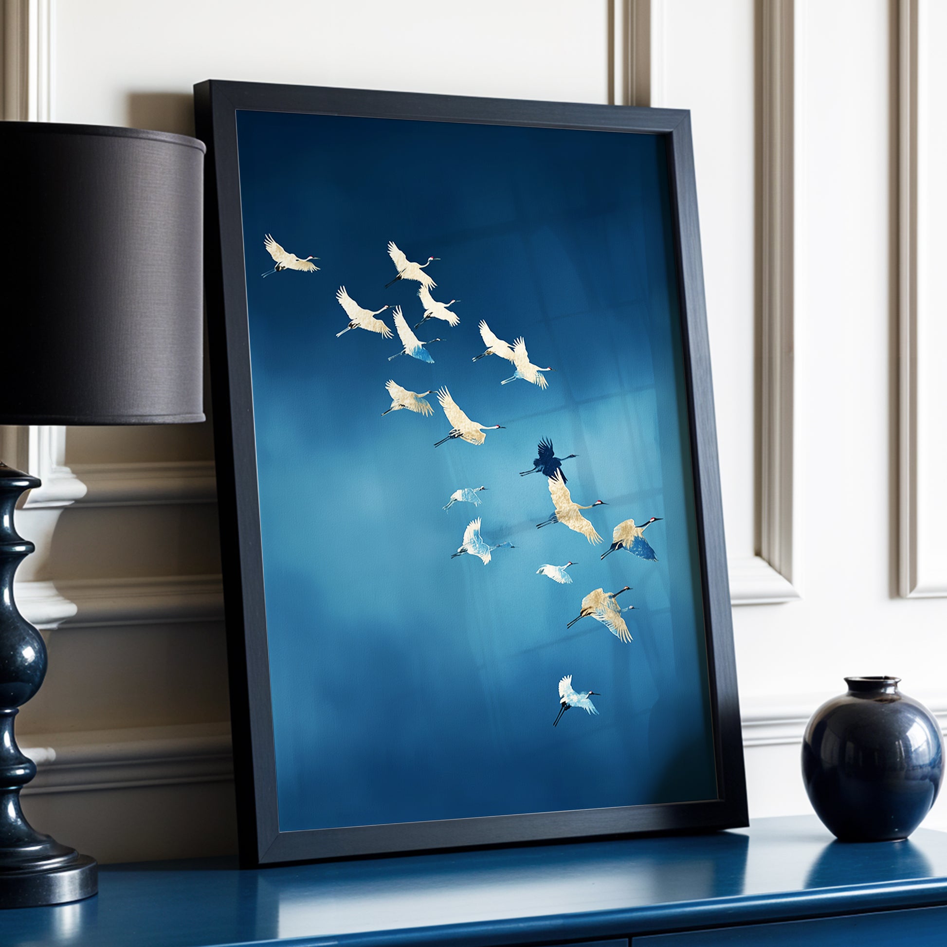 A framed art print featuring a flock of white cranes flying diagonally across a deep blue background, styled on a blue console table next to a black lamp and a small black vase.