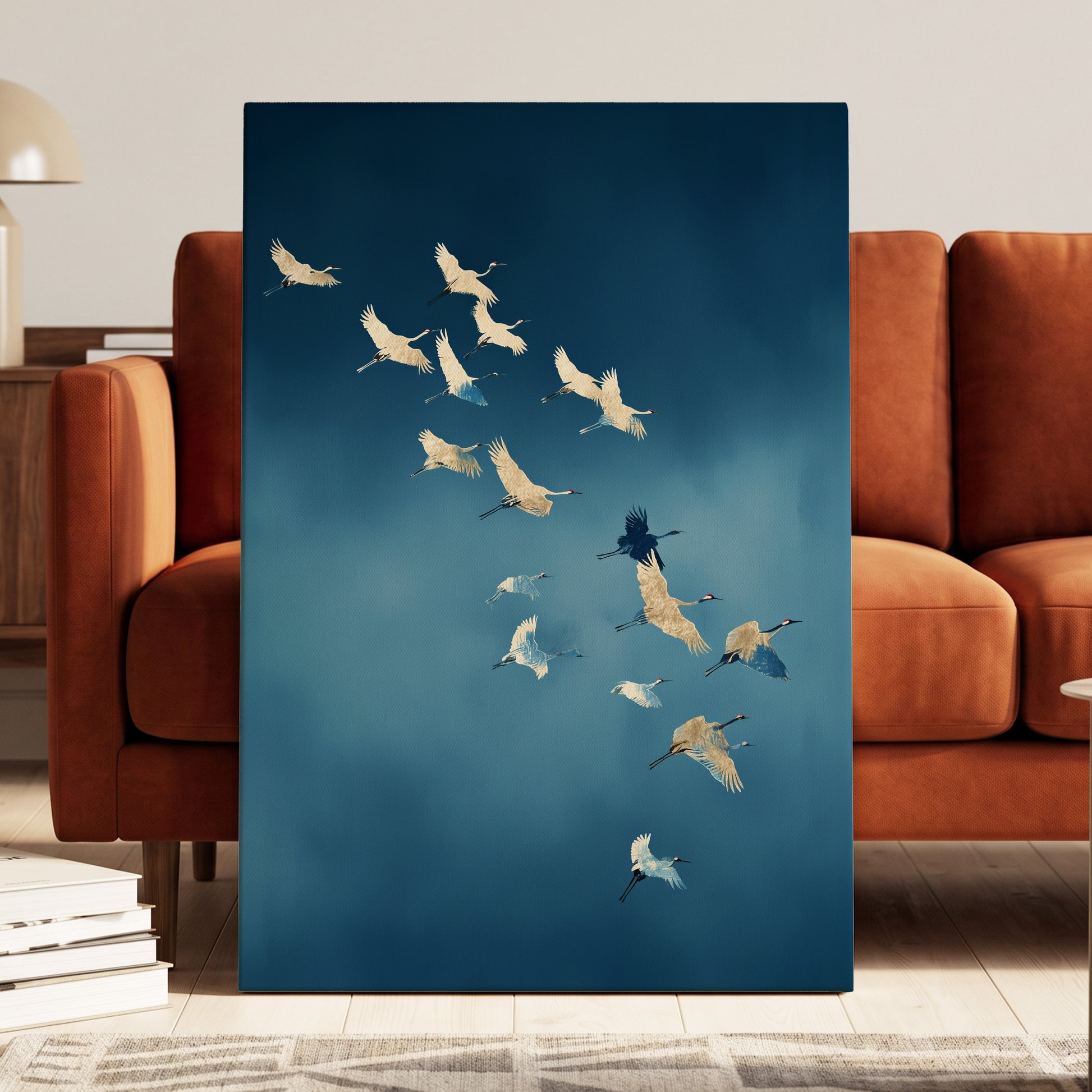 A vertical canvas wall art print featuring a flock of white cranes flying against a deep blue sky, styled leaning against a modern rust-brown leather sofa in a living room setting.