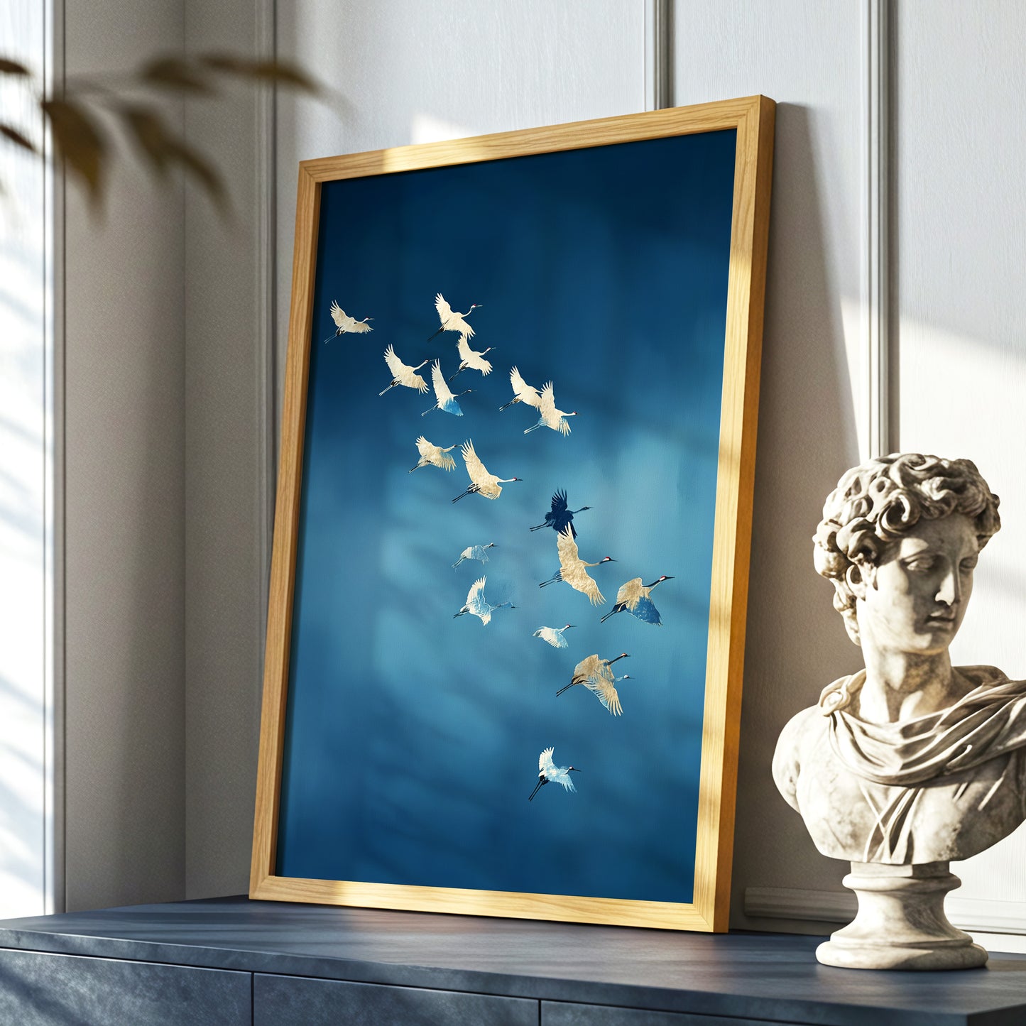 A framed art print leaning against a wall on a console table, featuring a flock of white and light blue cranes on a deep blue background. The print has a gold frame and is styled next to a classical bust sculpture.