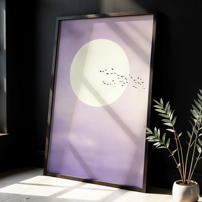 A framed minimalist art print leaning against a dark wall in a styled interior. The print features a large cream moon and a small flock of birds on a soft lavender-to-white gradient background.