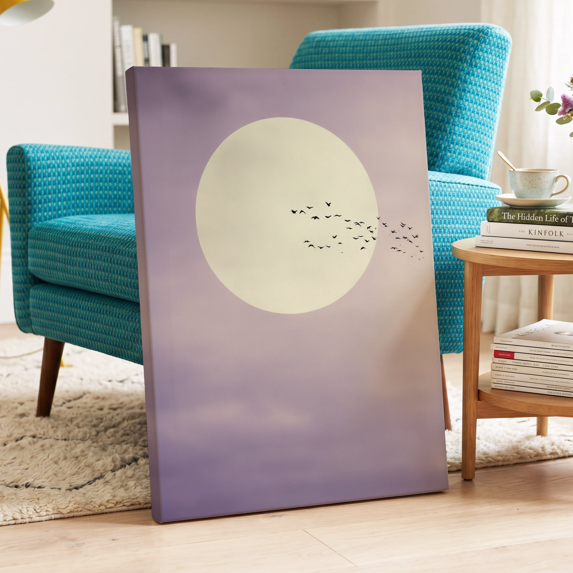 A vertical canvas wall art print leaning against a turquoise armchair in a bright living room. The artwork shows a large pale cream moon with a small flock of black birds against a soft lavender-to-purple sky.