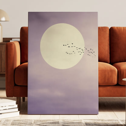 A vertical canvas wall art print leaning against a rust-brown sofa in a modern living room. The artwork features a large cream-colored full moon against a soft lavender-purple sky with a small flock of black birds flying from the right side.