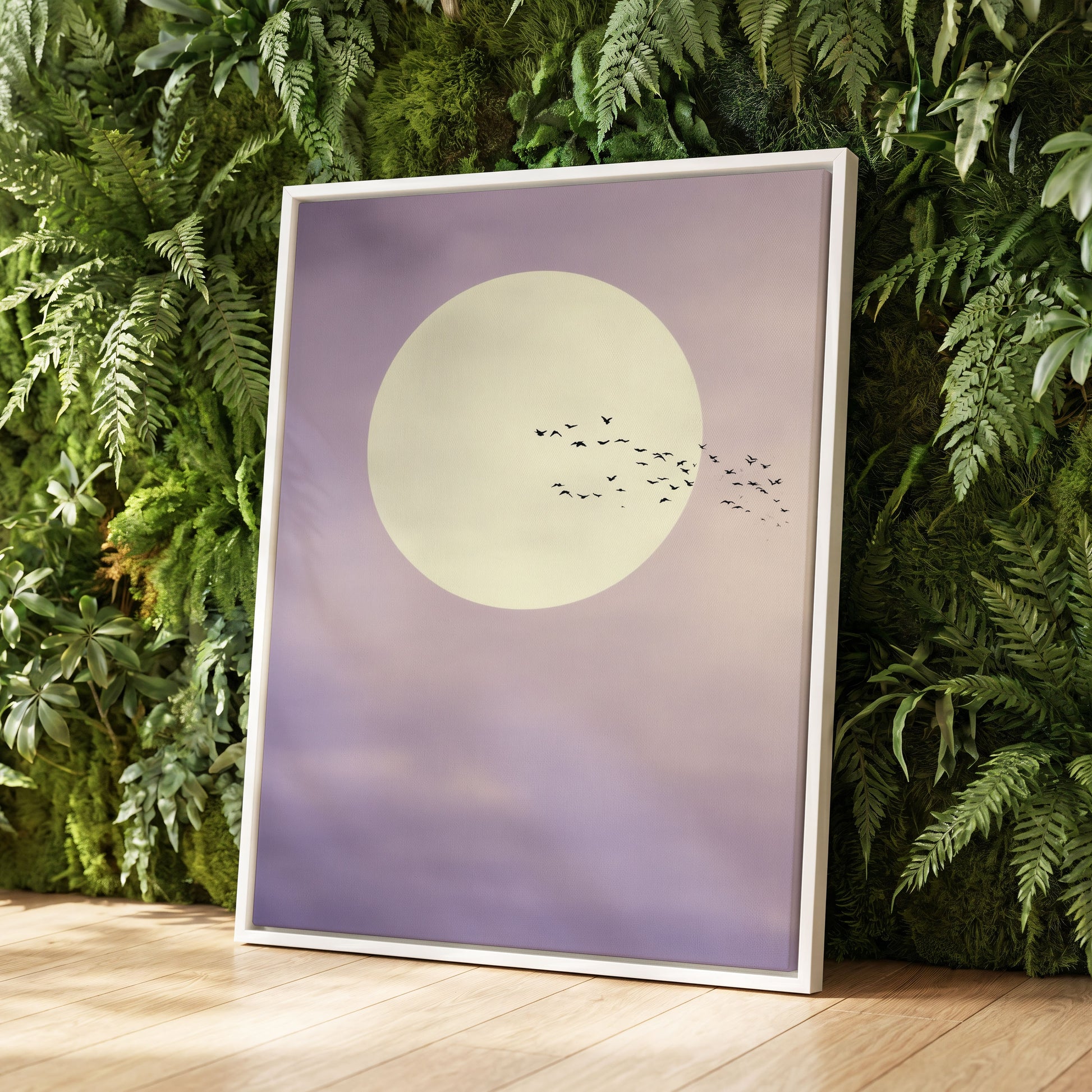 A framed art print featuring a large cream-colored moon and a flock of birds against a soft lavender-purple sky, leaning against a green fern wall on a light wood floor.