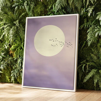 A framed art print featuring a large cream-colored moon and a flock of birds against a soft lavender-purple sky, leaning against a green fern wall on a light wood floor.