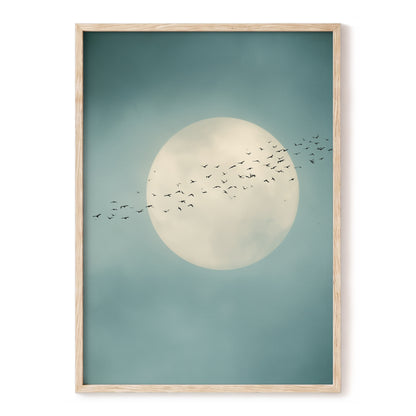 Framed wall art featuring a large cream moon and a flock of birds against a muted teal sky, shown in a light natural wood frame on a white background.