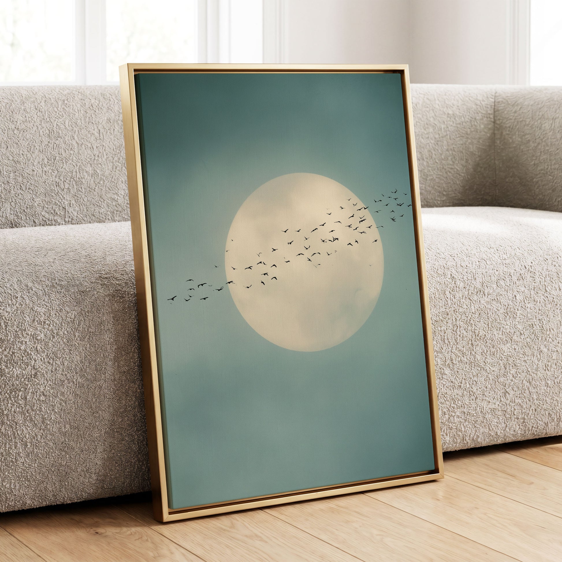 A framed art print leaning against a light gray textured sofa on a wooden floor. The print features a large cream-colored full moon with a flock of small black birds flying across a muted teal-blue sky, enclosed in a thin gold frame.