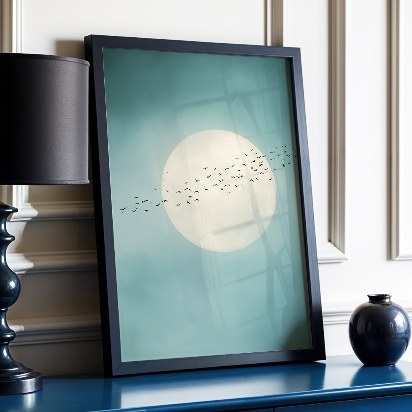 A framed art print leaning on a blue console table in a styled interior. The print features a large cream moon and a flock of birds against a muted teal background, enclosed in a black frame.