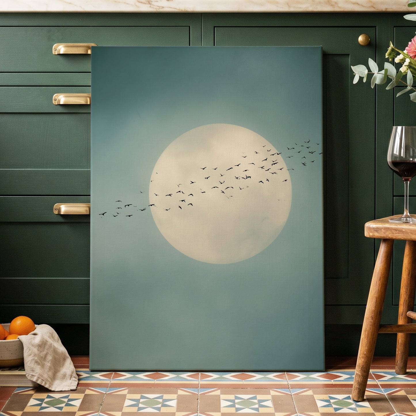 A vertical canvas wall art print featuring a large cream-colored full moon and a flock of black birds against a muted teal-blue sky, displayed leaning against dark green kitchen cabinets in a styled home interior.