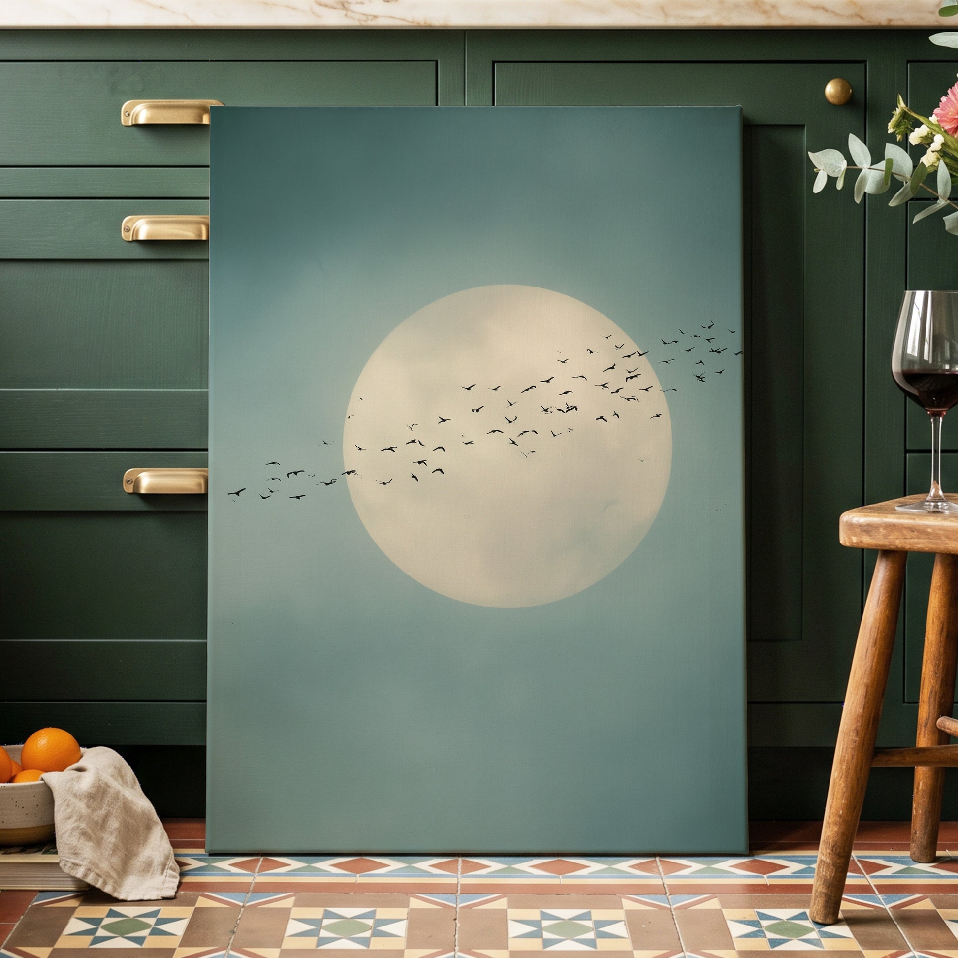 A vertical canvas wall art print featuring a large cream-colored full moon and a flock of black birds against a muted teal-blue sky, displayed leaning against dark green kitchen cabinets in a styled home interior.