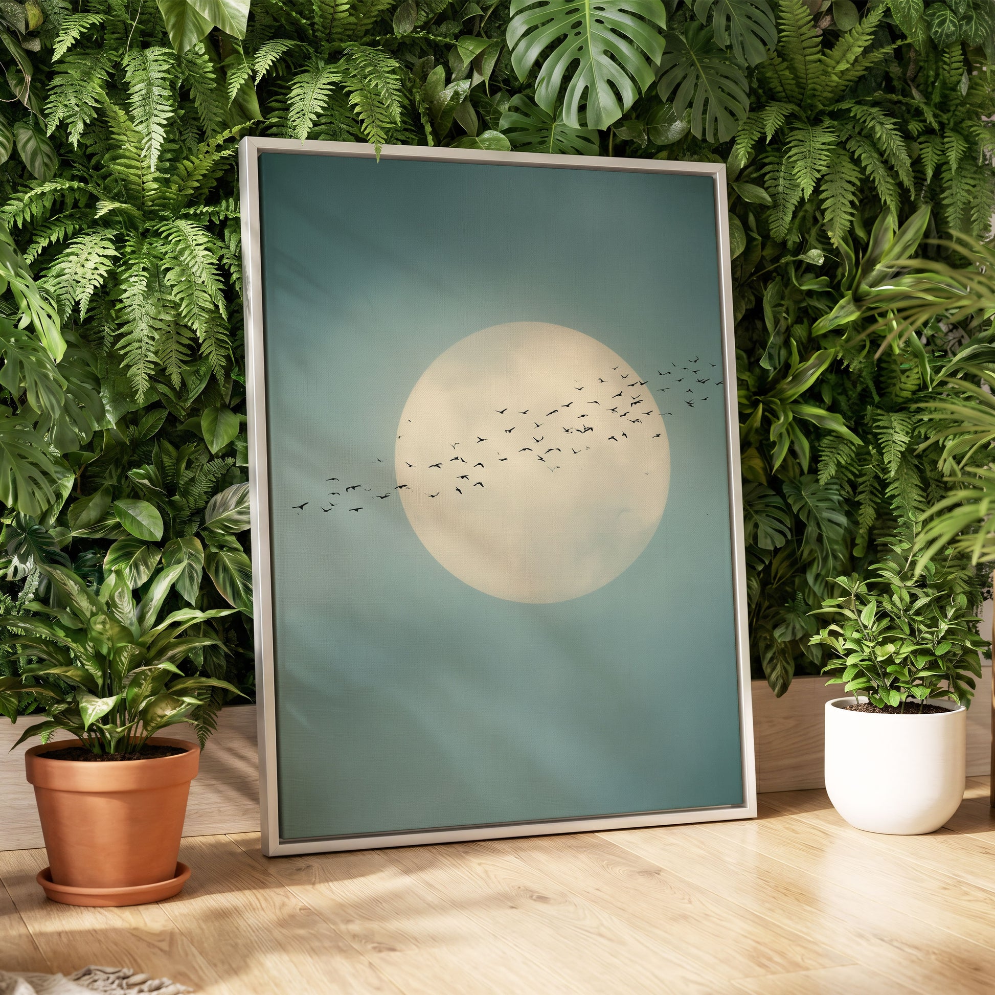 A framed art print featuring a large cream-colored moon with a flock of birds flying across a teal-blue sky, styled leaning against a wall of green houseplants on a light wood floor.