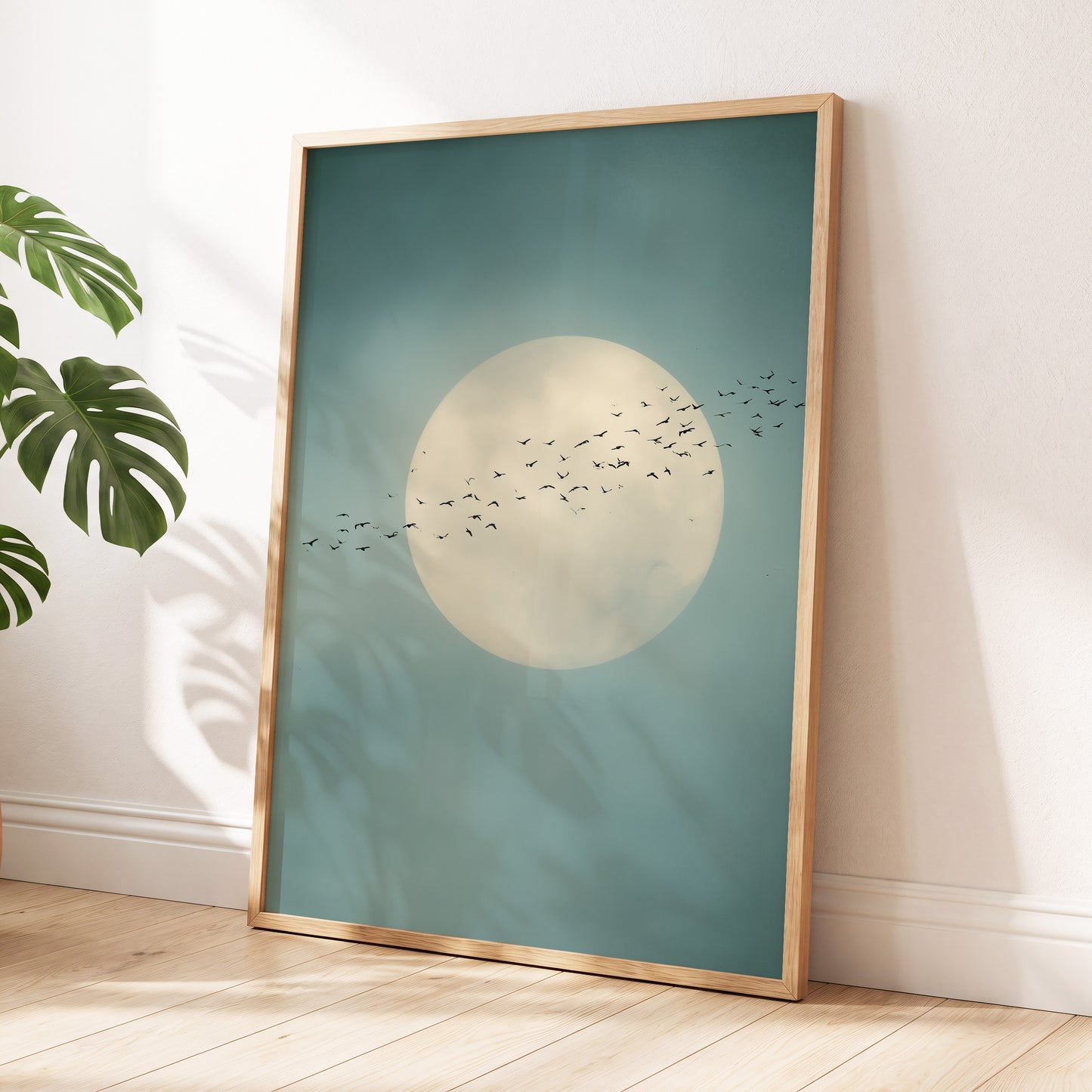 A framed art print leaning against a white wall on a light wood floor, featuring a large cream moon and a flock of birds over a muted teal-blue sky, with a gold-toned frame and a green plant leaf visible at the left edge.