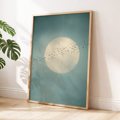 A framed art print leaning against a white wall on a light wood floor, featuring a large cream moon and a flock of birds over a muted teal-blue sky, with a gold-toned frame and a green plant leaf visible at the left edge.