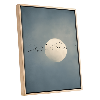 Framed wall art featuring a pale full moon centered against a muted blue-gray cloudy sky, with small black bird silhouettes flying around it. The artwork is shown in a light natural wood frame on a plain white background.