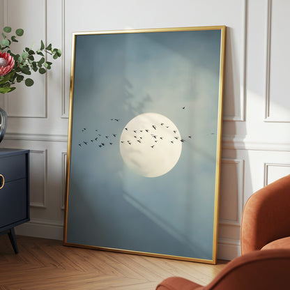 A large framed wall art print leaning against a white paneled wall in a styled room. The artwork shows a pale moon with small black birds flying across a soft blue-gray sky.