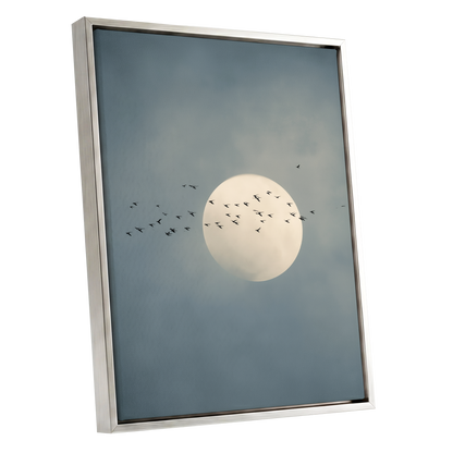 Framed wall art featuring a pale full moon against a muted blue-gray cloudy sky, with small black bird silhouettes flying around it. The artwork is shown in a slim metallic frame on a plain white background.