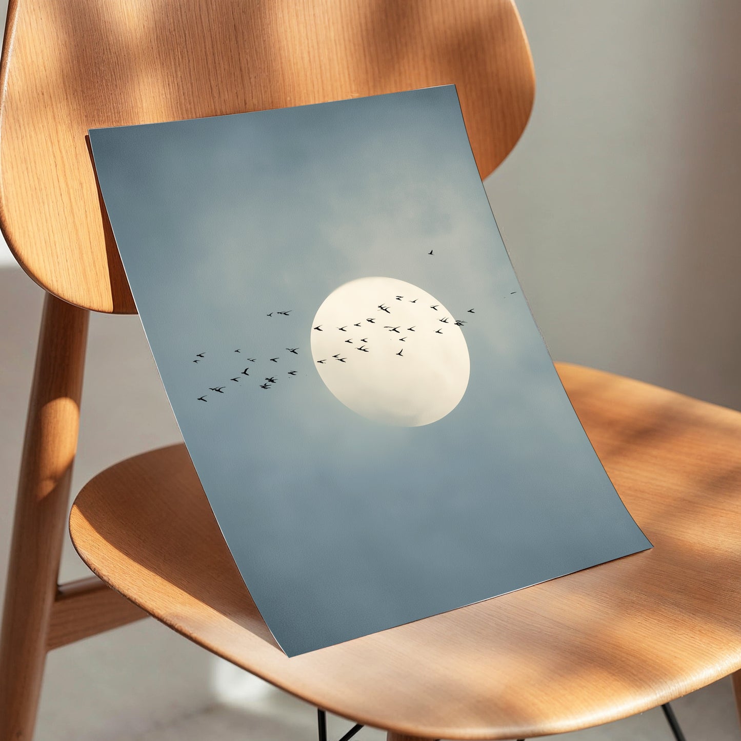 A square art print depicting a large white full moon surrounded by a flock of small black birds against a soft blue-gray sky, displayed on a light brown wooden chair.