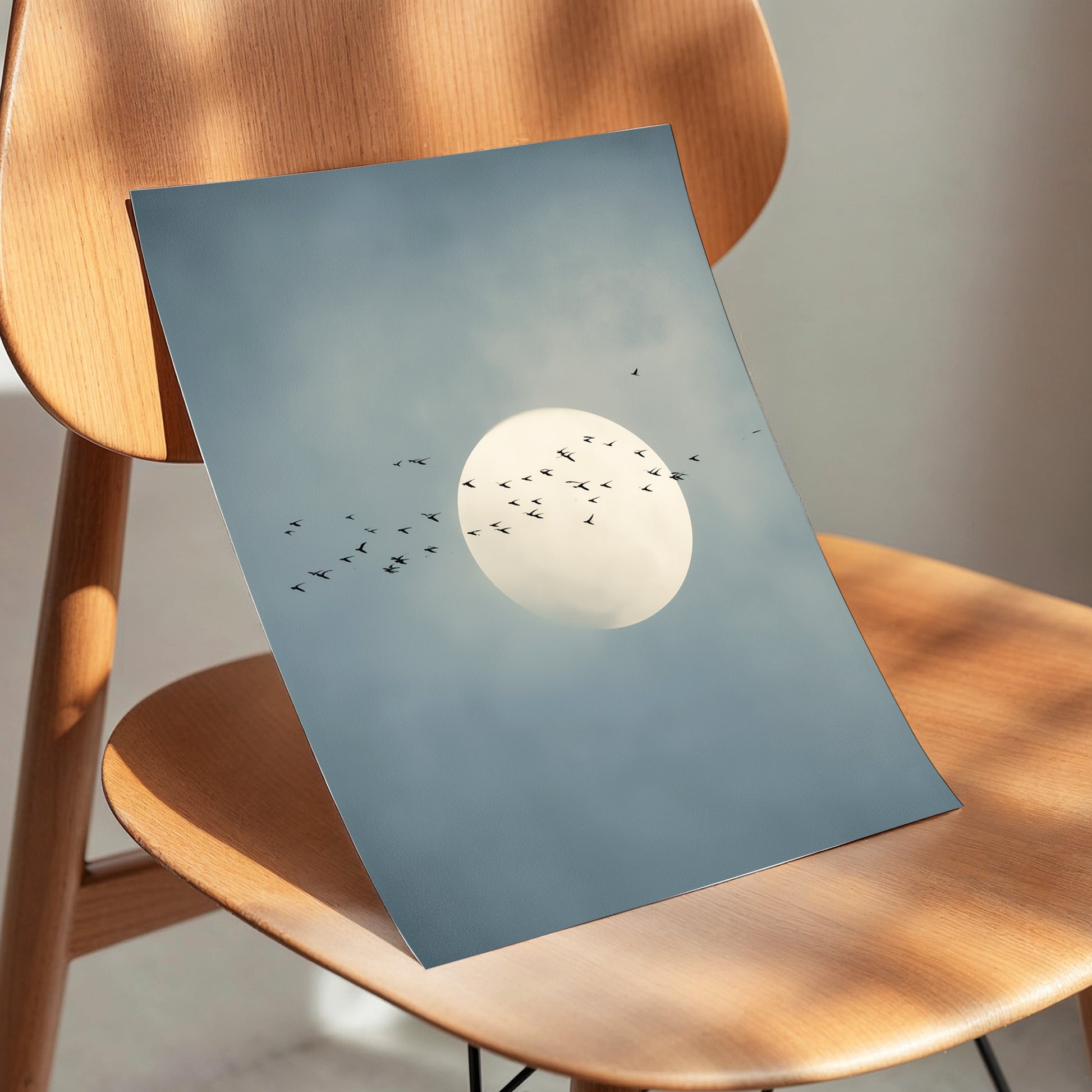 A square art print depicting a large white full moon surrounded by a flock of small black birds against a soft blue-gray sky, displayed on a light brown wooden chair.