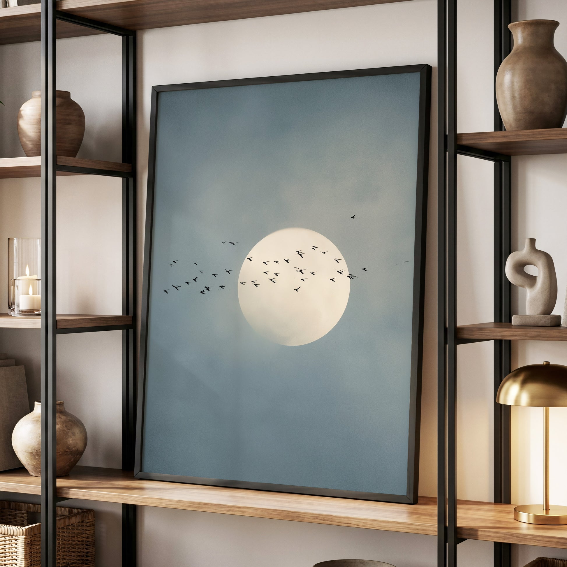 A framed art print featuring a large white moon and small flying birds against a muted blue-gray sky, displayed on a wooden shelf within a modern black metal shelving unit.