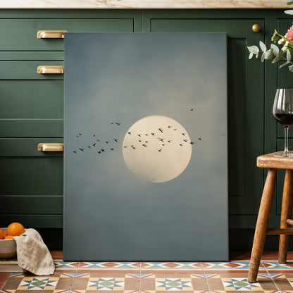 A vertical canvas wall art print featuring a large cream-colored full moon and a flock of small black birds against a muted slate-blue sky, displayed leaning against dark green kitchen cabinets in a styled interior setting.
