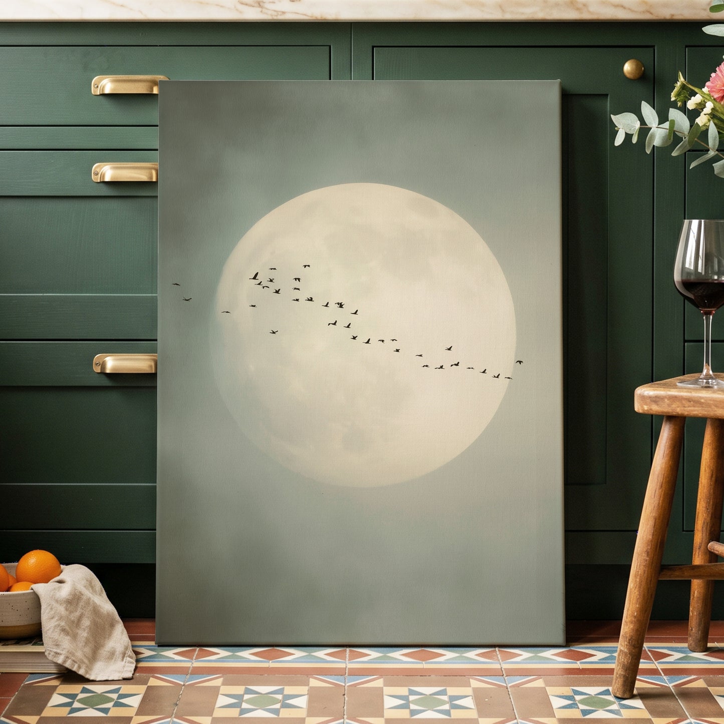 A large vertical canvas print leaning against dark green kitchen cabinets. The artwork depicts a large pale full moon with a flock of small birds flying across it.