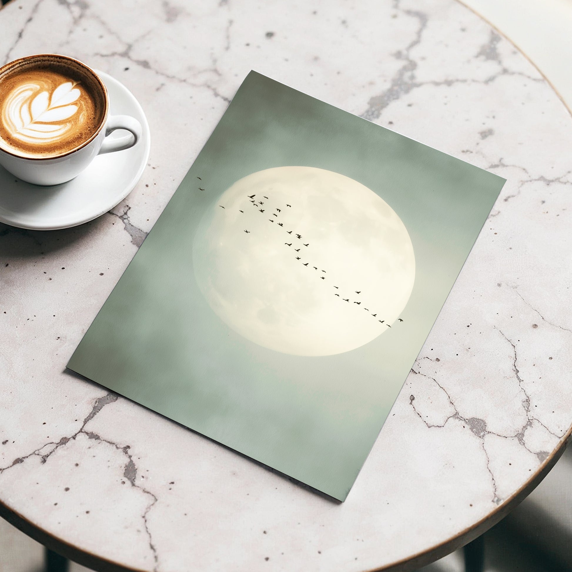 A square art print featuring a large pale moon with a line of small birds across it, styled on a round marble table next to a cup of latte.