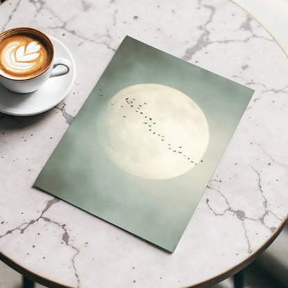 A square art print featuring a large pale moon with a line of small birds across it, styled on a round marble table next to a cup of latte.