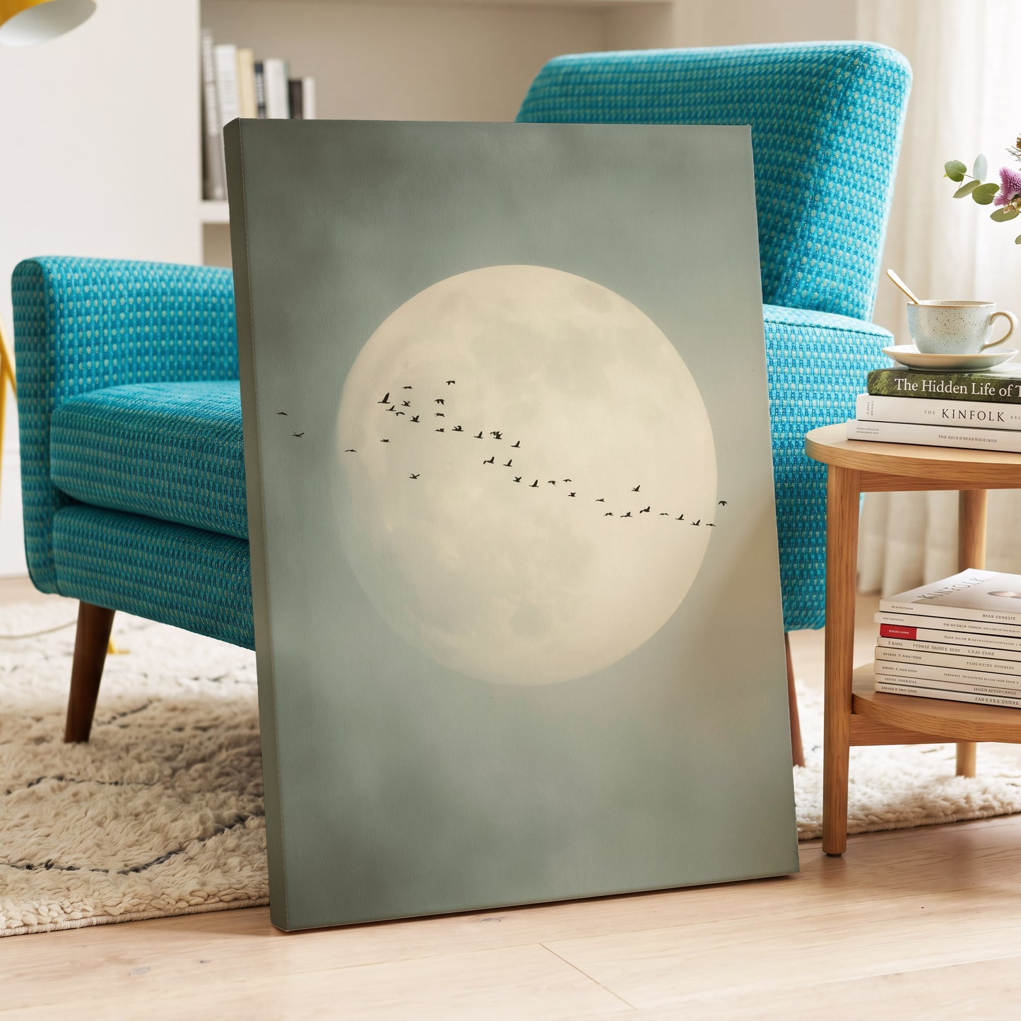 A vertical canvas wall art print leaning against a turquoise armchair in a styled living room. The artwork depicts a large pale full moon with a flock of small black birds flying across it on a muted sage-gray background.
