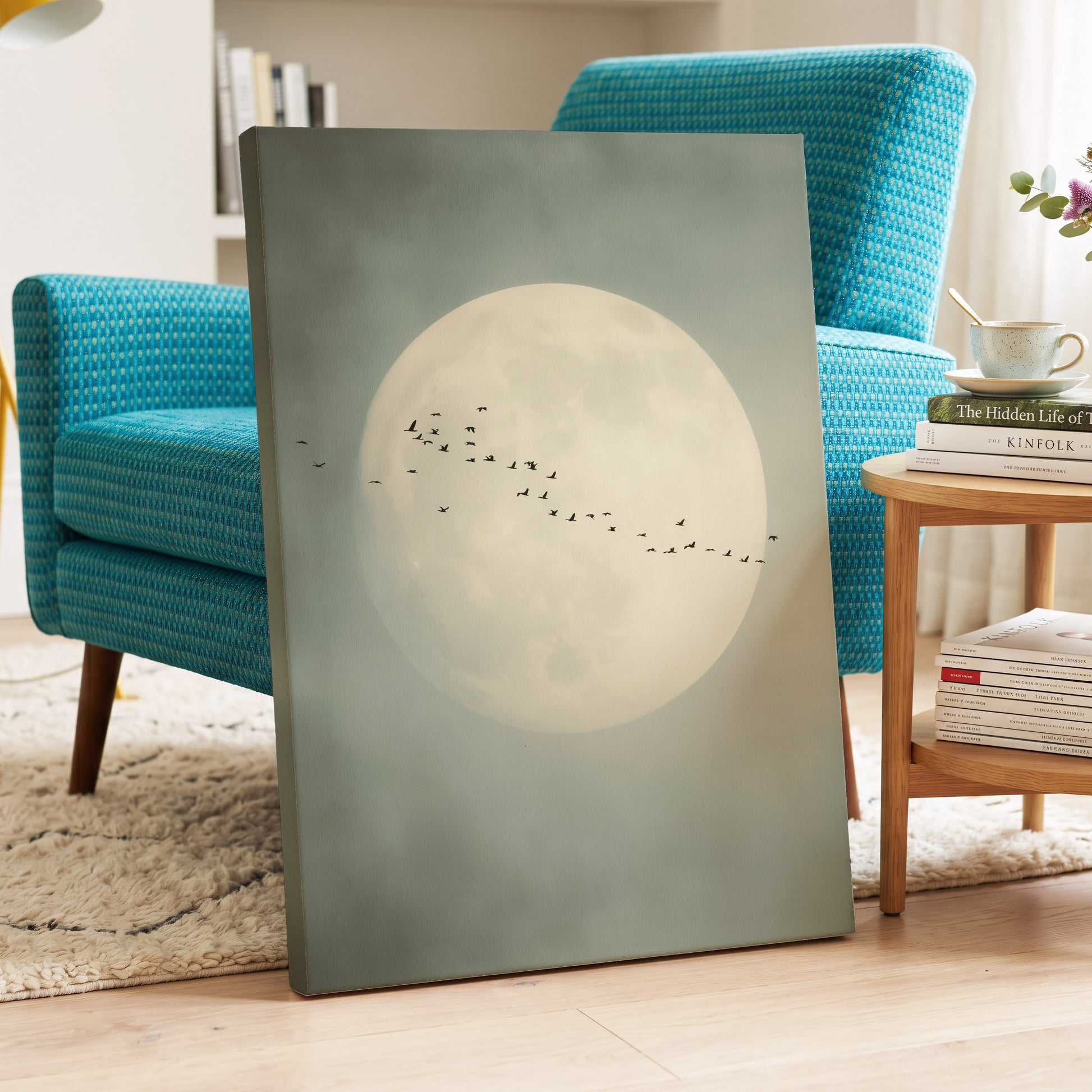 A vertical canvas wall art print leaning against a turquoise armchair in a styled living room. The artwork depicts a large pale full moon with a flock of small black birds flying across it on a muted sage-gray background.