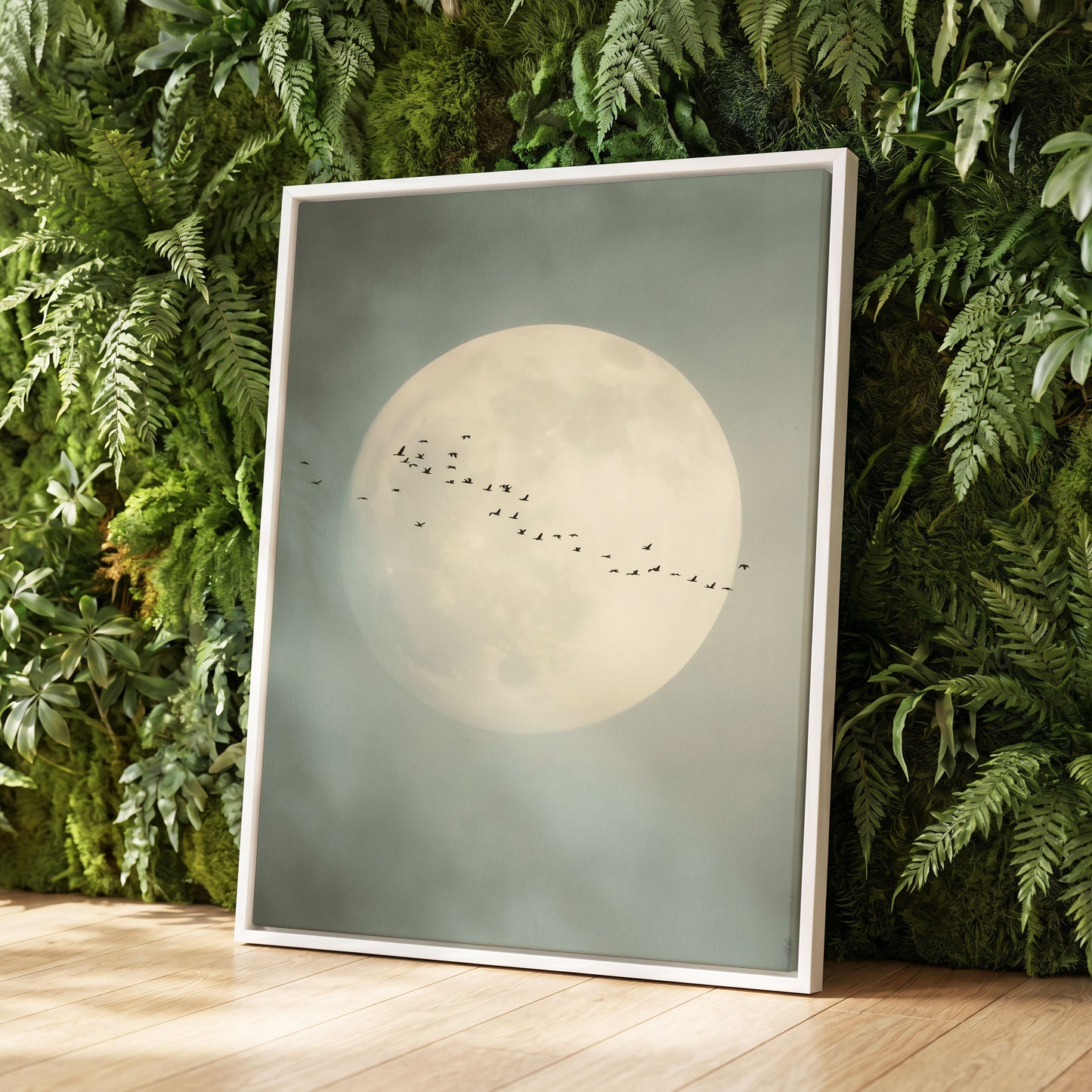A framed art print featuring a large pale moon and a flock of small birds against a misty gray-green sky, leaning against a lush green fern wall on a light wood floor.