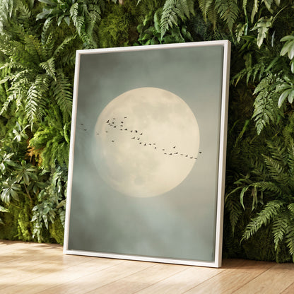 A framed art print featuring a large pale moon and a flock of small birds against a misty gray-green sky, leaning against a lush green fern wall on a light wood floor.