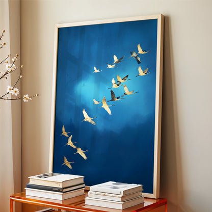 Large framed wall art featuring a flock of cranes flying across a deep blue background, displayed leaning against a beige wall above a small wooden table with stacked books.