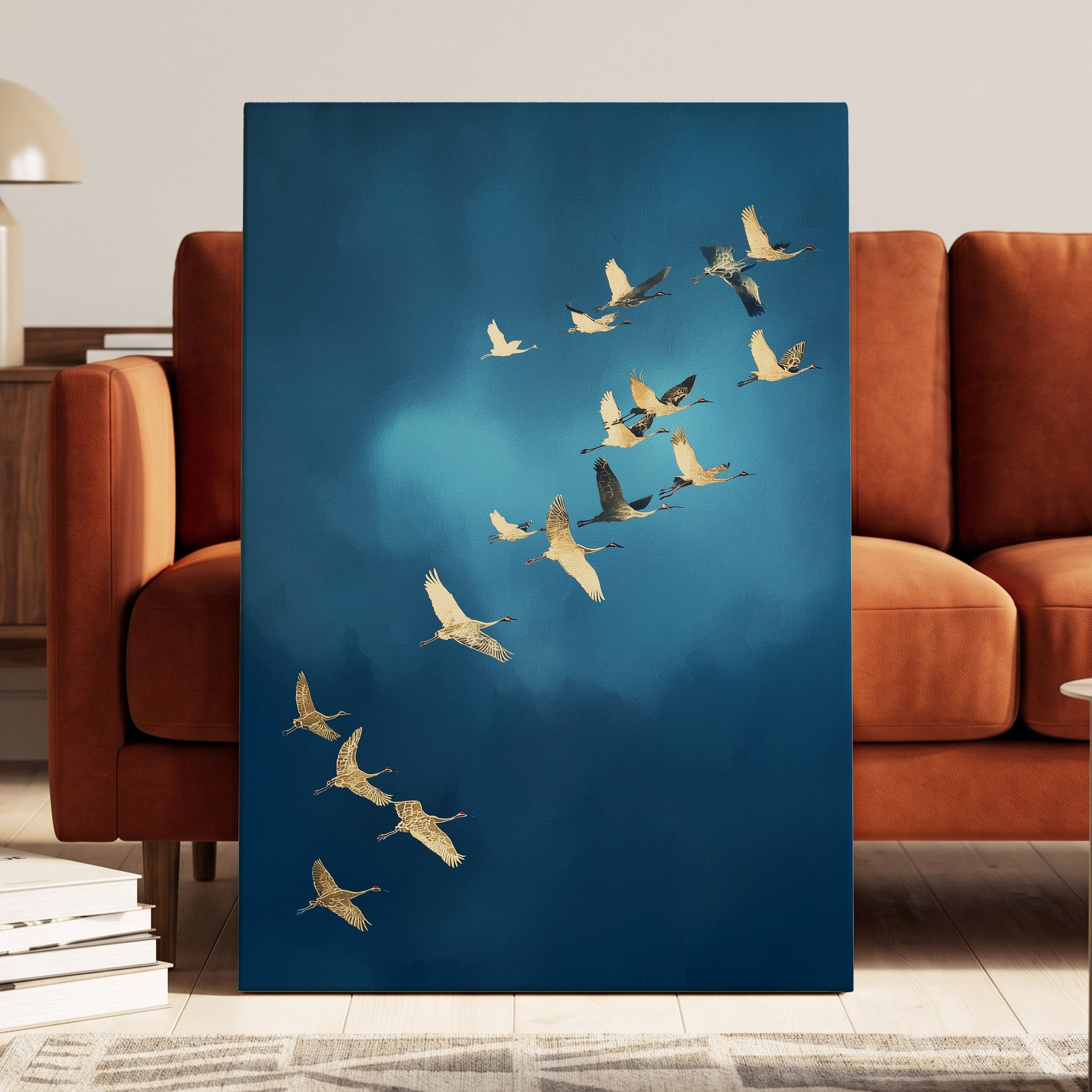 A vertical canvas wall art print depicting a flock of cranes flying across a deep blue sky, styled leaning against a modern rust-brown leather sofa in a living room setting.