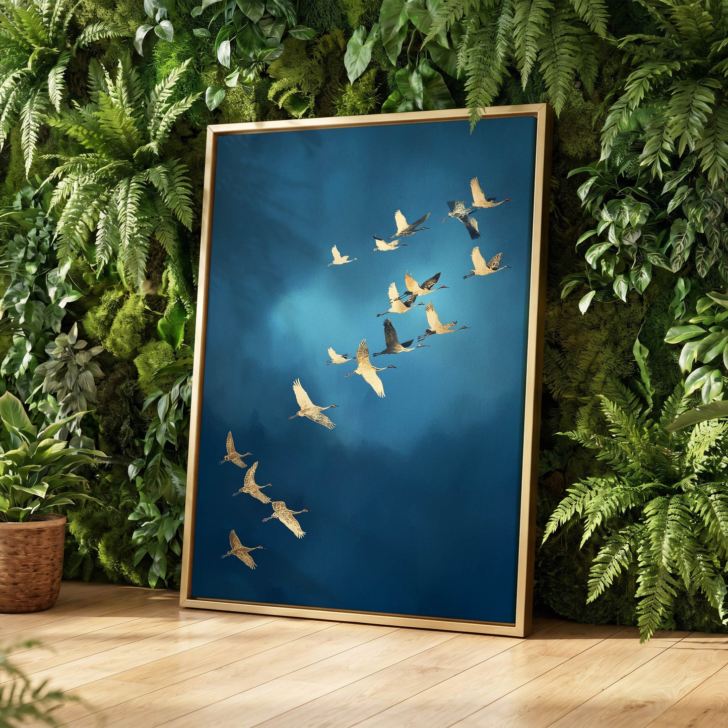 A framed wall art print leaning against a lush green moss wall on a light wood floor. The artwork shows a flock of birds flying across a deep blue sky, and the frame is a thin gold tone.