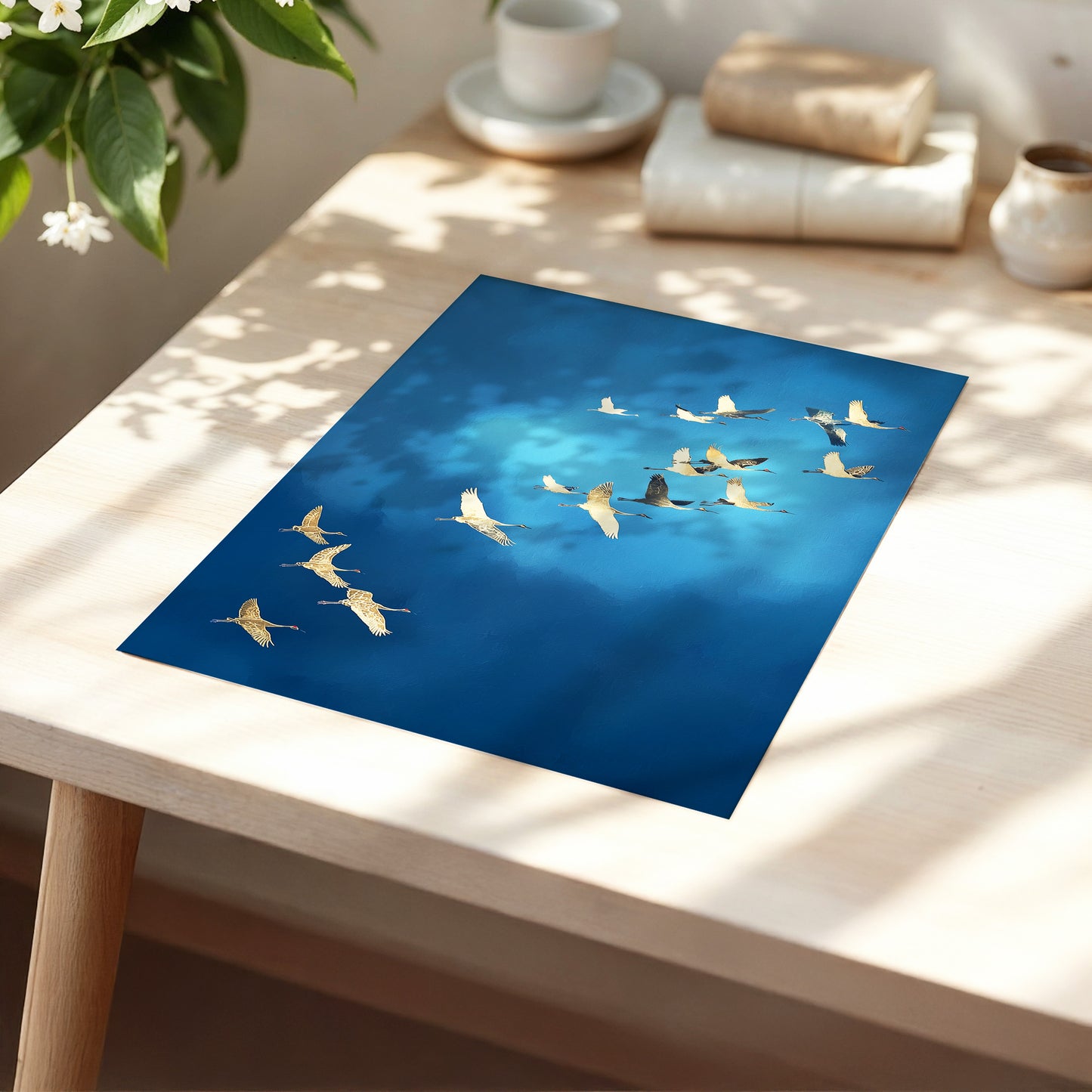 A square art print featuring a flock of golden cranes flying against a deep blue sky, displayed on a light wood table in a sunlit, styled room.