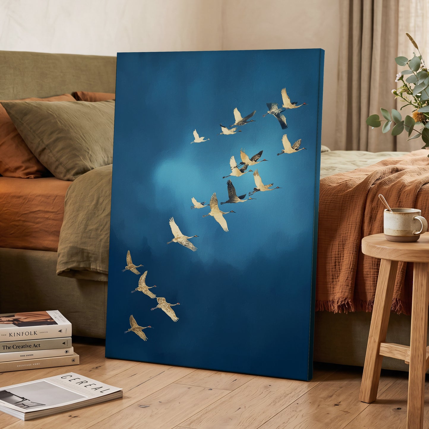 A vertical canvas wall art print leaning against a bed in a styled bedroom. The artwork depicts a flock of cranes flying diagonally across a deep blue sky with a soft light glow.