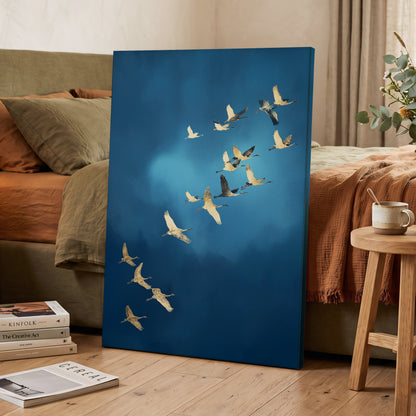 A vertical canvas wall art print leaning against a bed in a styled bedroom. The artwork depicts a flock of cranes flying diagonally across a deep blue sky with a soft light glow.
