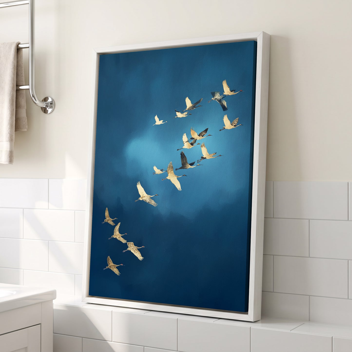 A framed vertical canvas print of a flock of cranes flying against a deep blue sky, leaning against a bathroom wall with white subway tiles.