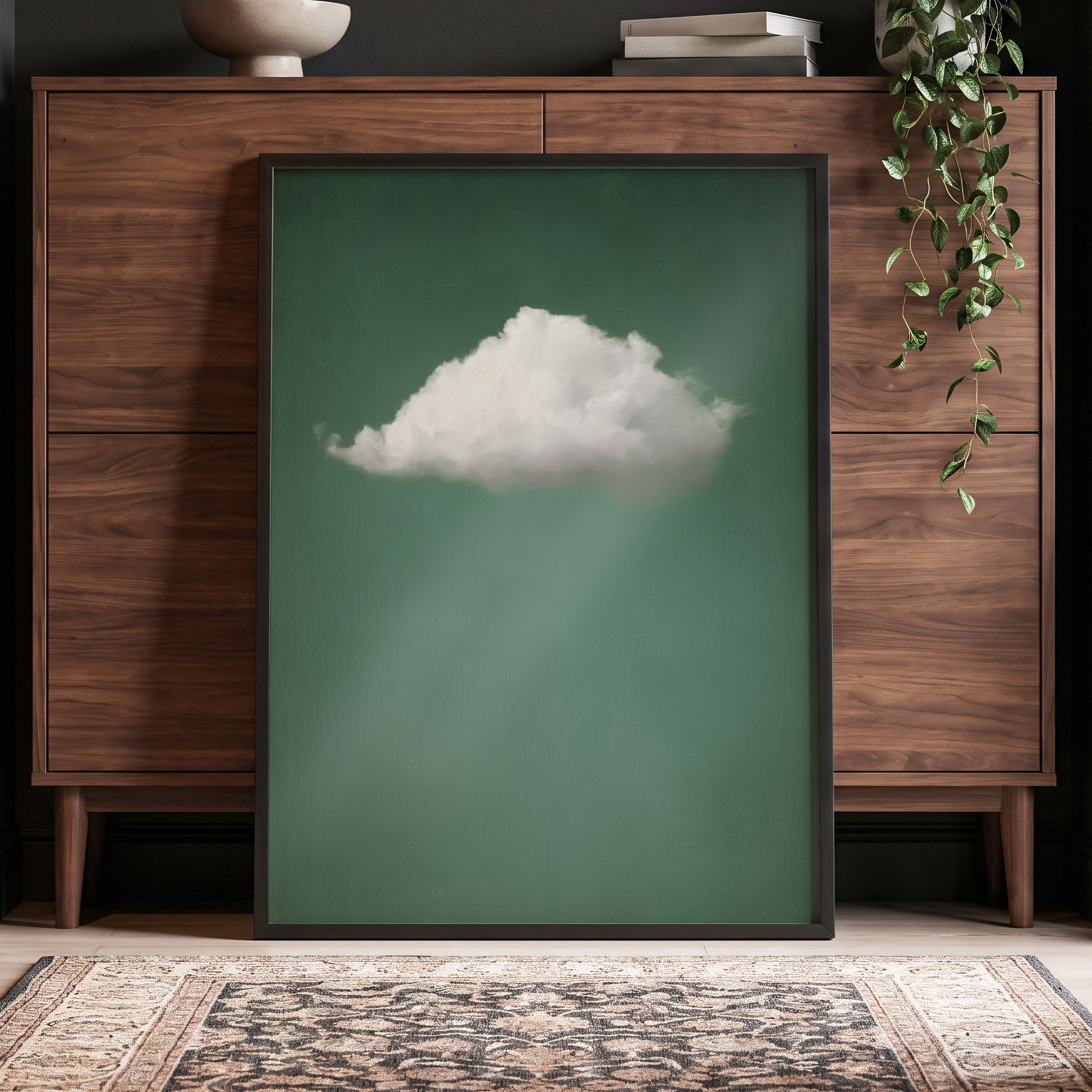 A framed art print featuring a single white cloud against a muted green background, leaning against a mid-century modern wooden cabinet in a styled room setting.