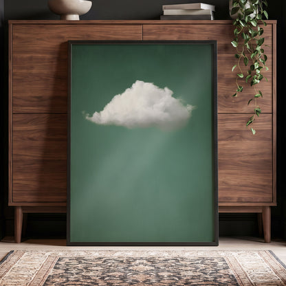 A framed art print featuring a single white cloud against a muted green background, leaning against a mid-century modern wooden cabinet in a styled room setting.