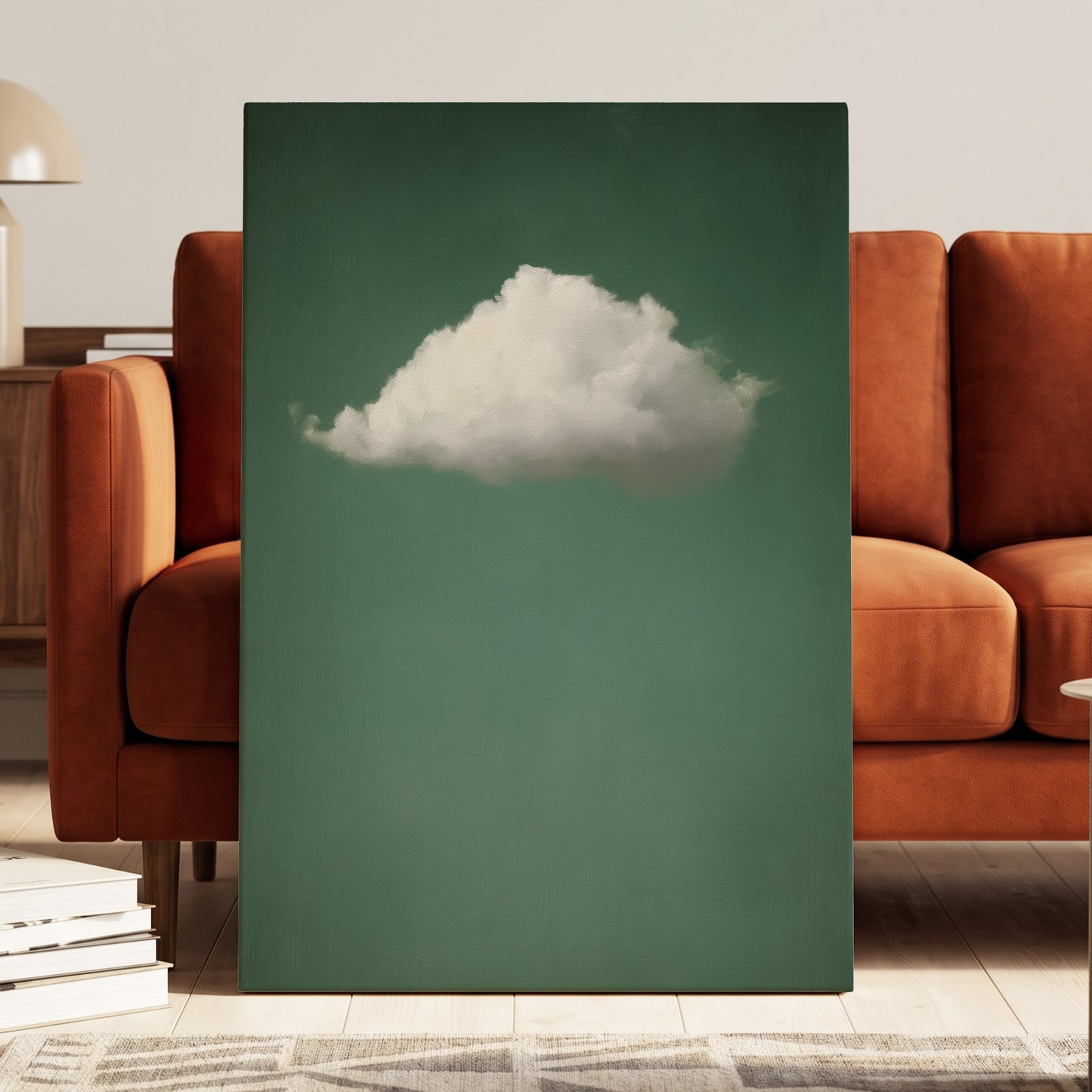 A vertical canvas wall art print featuring a white cloud against a muted teal-green background, styled leaning against a rust-orange sofa in a modern living room setting.