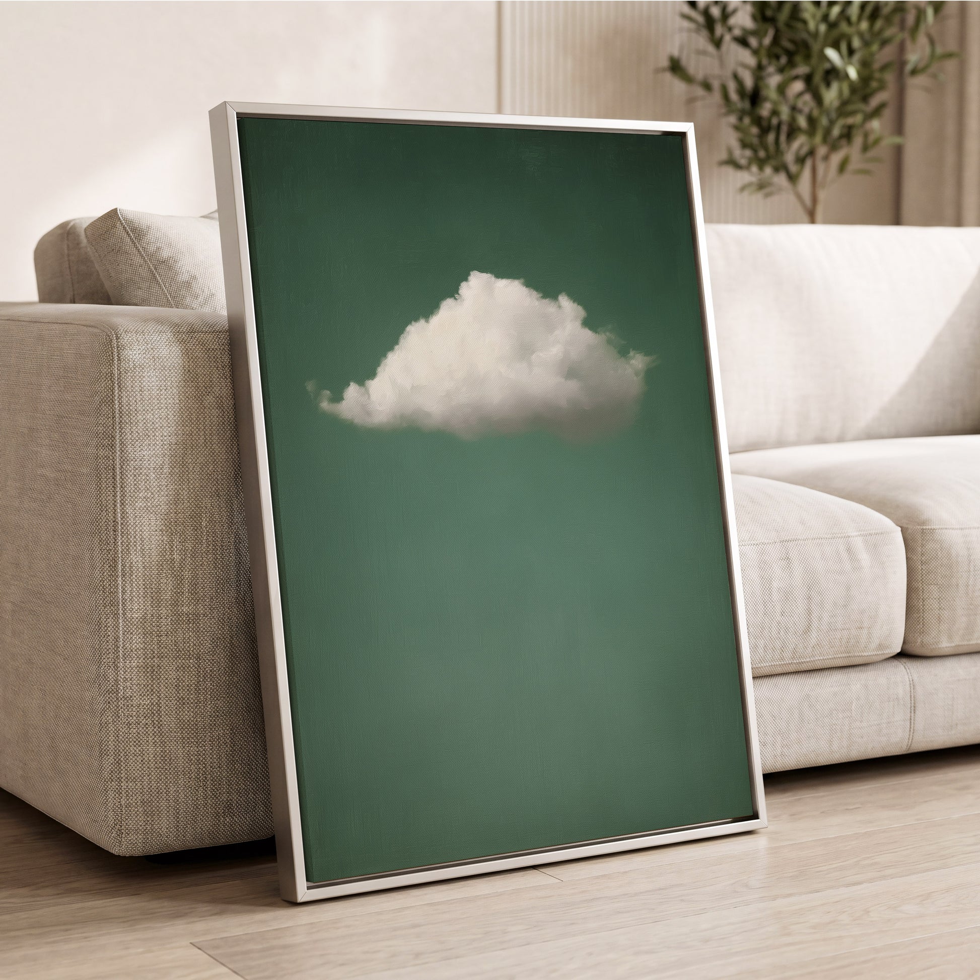 A framed wall art print leaning against a light beige sofa in a living room. The print shows a single white cloud centered on a muted green background, with a thin metallic frame.